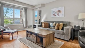 A living room with a wooden coffee table, grey sofa, and a window with curtains. - The Dragonfly | Pool/Hot Tub | Lake Access | Kayak (Invermere)