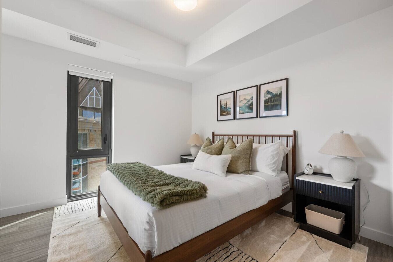 A bedroom with a large bed, a nightstand with a lamp, a window with a view of a building, and three framed pictures on the wall.
