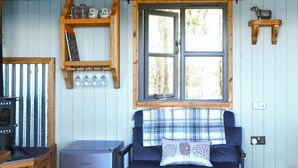 A cozy room with a blue sofa, a small refrigerator, a window with a view of trees, and wooden shelves with mugs and glasses. - 1 bedroom accommodation in Castle Douglas (Castle Douglas)