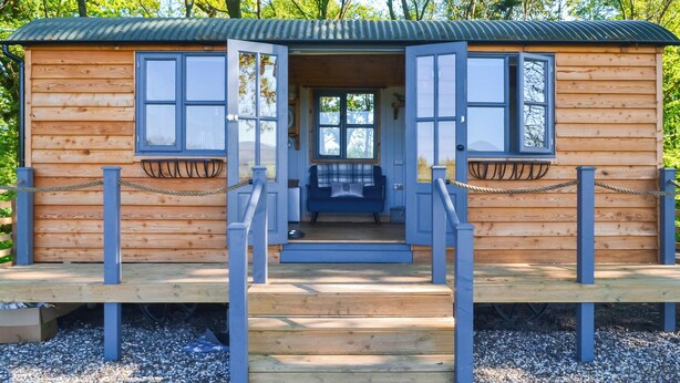 A wooden cabin with a porch, two windows, and a door leading inside. - 1 bedroom accommodation in Castle Douglas (Castle Douglas)