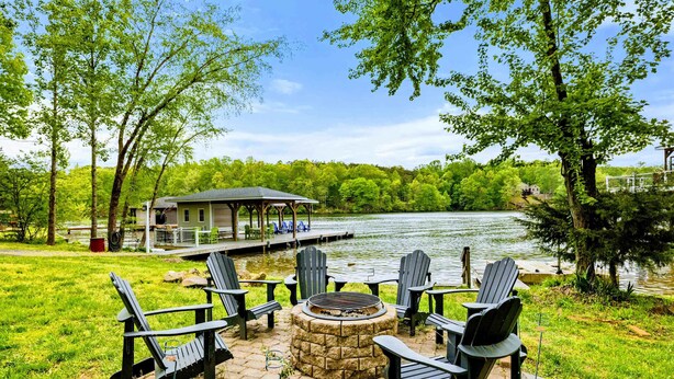 A lakeside deck with Adirondack chairs and a fire pit, overlooking a calm lake and a house with a covered deck.