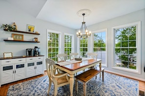 A dining area with a wooden table, white chairs, a bench, and a chandelier. There are large windows with a view of greenery outside.