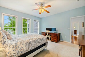 A bedroom with a bed, a desk, a chair, a ceiling fan, and large windows.