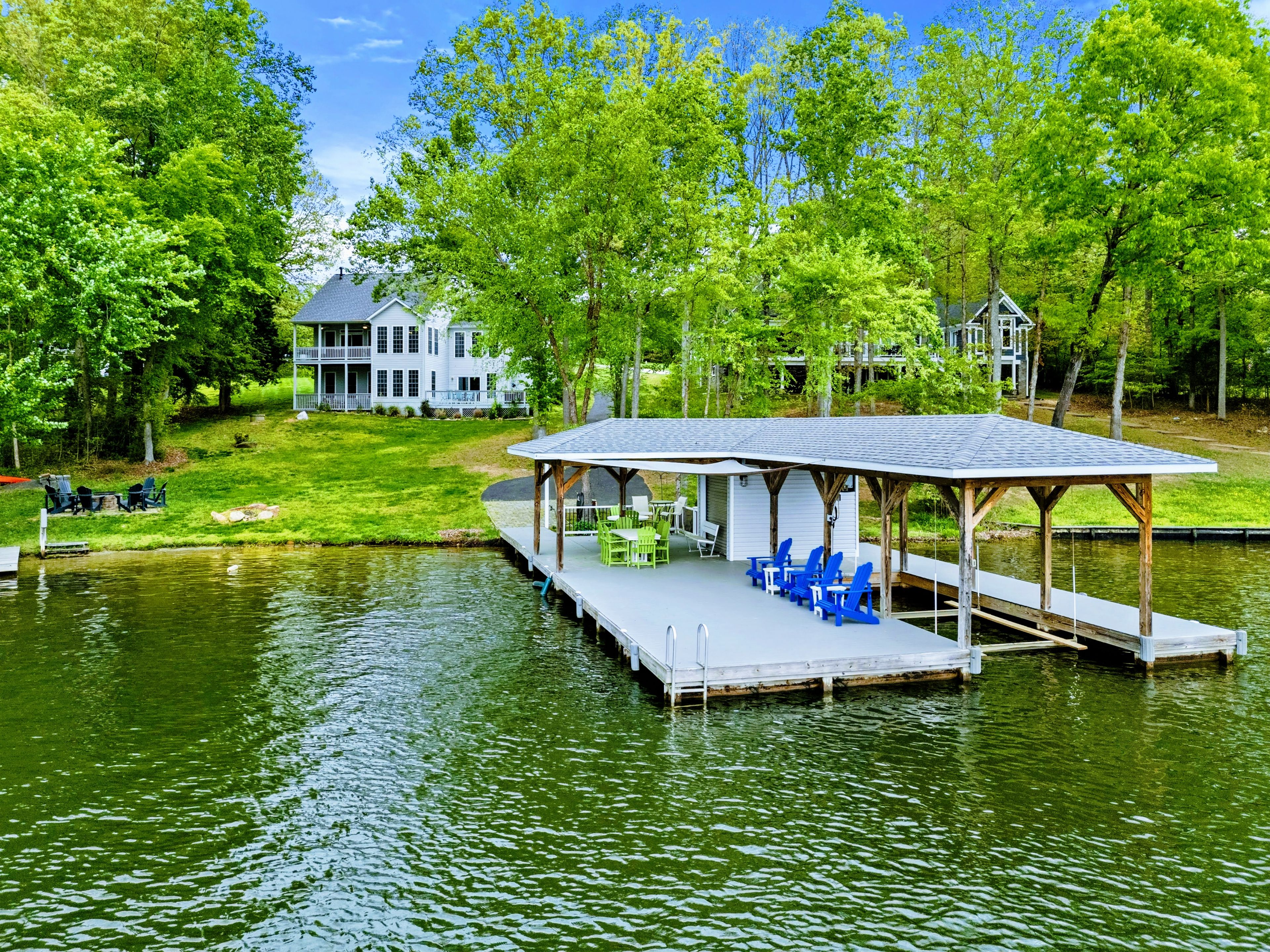 A lakeside property with a dock, outdoor seating, and a large house in the background.