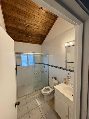 Kylpyhuone, jossa on puinen katto, lasinen suihkusein ä, wc, pesuallas ja valoilla varustettu peili.