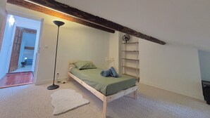 Une petite chambre avec un lit simple, un plafond en pente, un ventilateur sur pied, une table de chevet et une lampe sur pied.