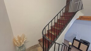Un escalier avec une rampe en bois et des balustrades en métal noir, menant à un palier où se trouvent une petite table et un vase de fleurs séchées.