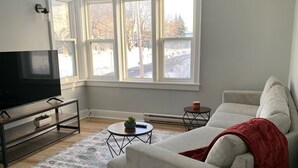 Ein Wohnzimmer mit einer Couch, einem Couchtisch, einem Fernseher auf einem Schrank und einem Fenster mit Blick auf Bäume und Gebäude.