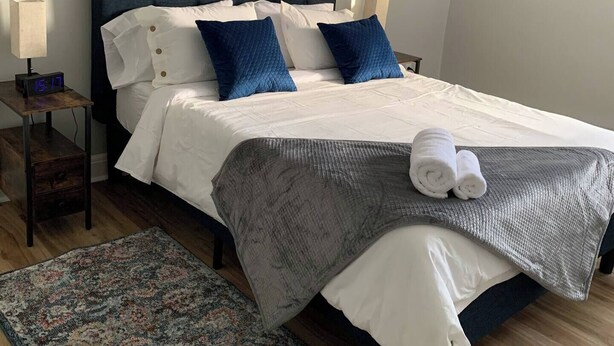 Ein ordentlich bezogenes Bett mit weißer und blauer Bettwäsche, einer grauen Decke und zusammengerollten Handtüchern an der Seite.