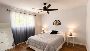 Um quarto com cama, duas mesas de cabeceira, um ventilador de teto, uma janela com cortinas e uma porta.