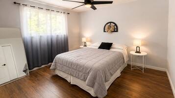 A bedroom with a bed, two bedside tables, a ceiling fan, a window with curtains, and a door.