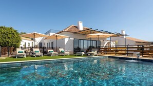 Une piscine entourée de chaises longues et de parasols, bordée d’un bâtiment blanc doté de grandes fenêtres et d’un toit recouvert de tuiles.
