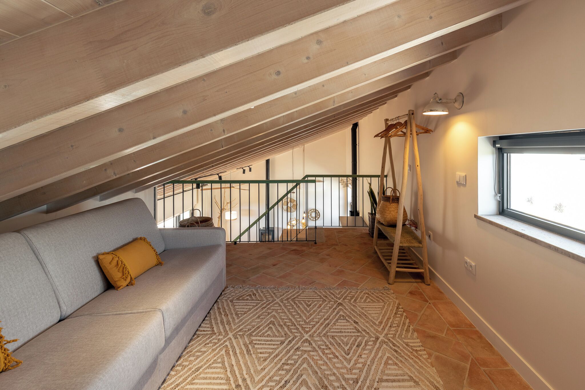 Dormitorio tipo loft con techo inclinado, sofá gris, alfombra estampada y ventana con vistas.