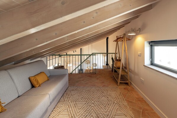 Une chambre à coucher aménagée dans un espace mansardé, avec un plafond en pente, un canapé gris, un tapis à motifs et une fenêtre offrant une vue dégagée.