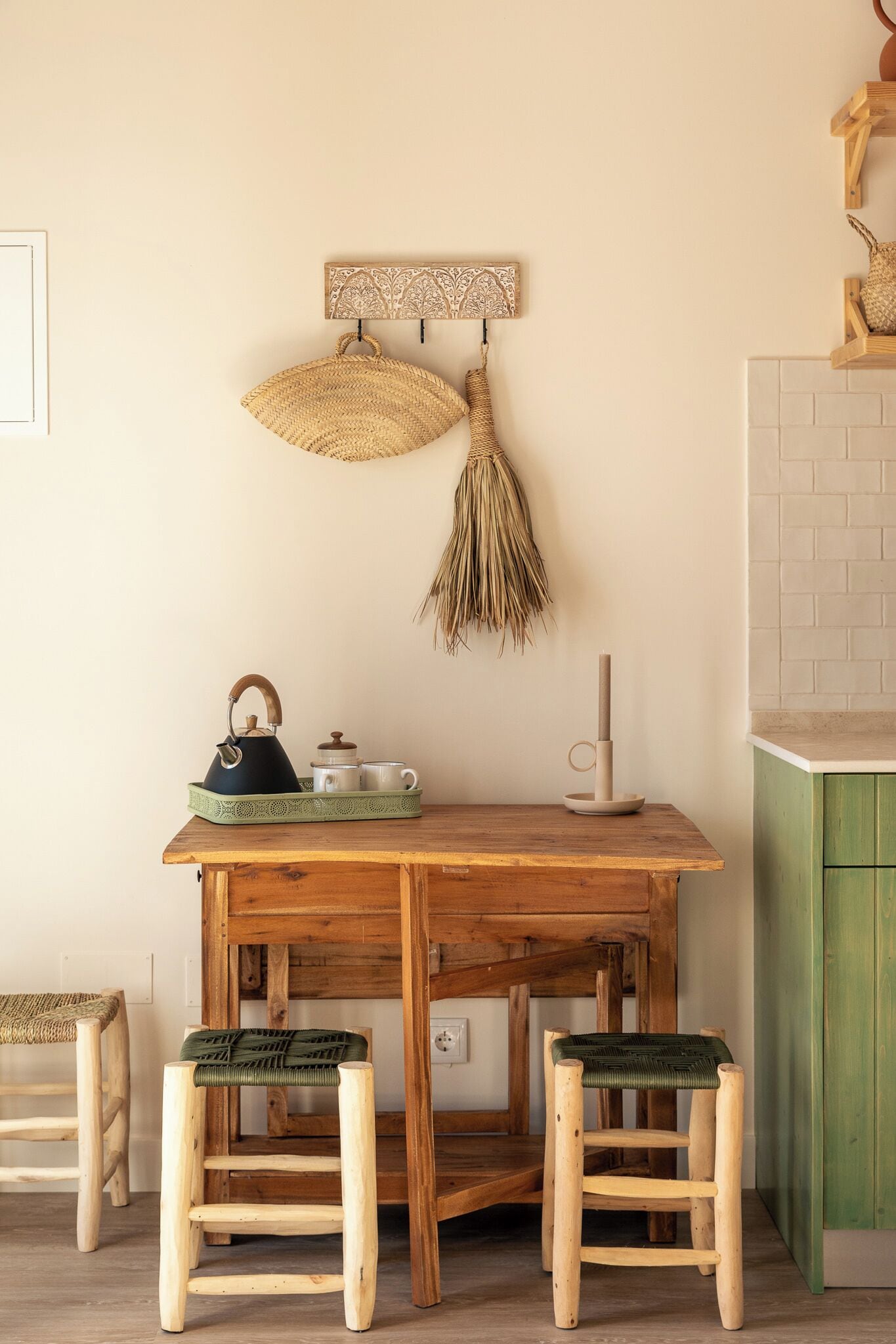 Een houten eettafel met twee krukken, een gevlochten mand, een theeservies en een decoratief object aan de muur.
