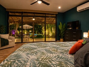 A bedroom with a bed, a TV, a dresser, and a view of a pool and garden through a sliding glass door.