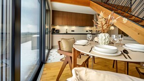 Et moderne spiseområde med et rundt bord dækket til to personer, træstole og udsigt til køkken og balkon.