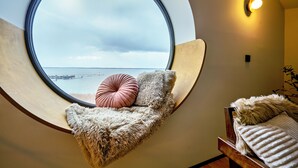 Een kamer met een rond raam met uitzicht op een wateroppervlak, een houten bankje met een zachte deken en een roze kussen, en een wandlamp.
