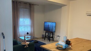 A living room with a wooden table, a television, a green sofa, and a vacuum cleaner. - Apartment 85m2 short term rental 1 month 2 months 3 months max available (Bourgoin-Jallieu)