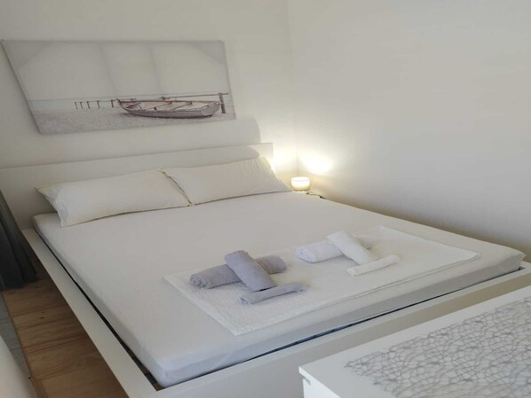 A neatly made bed with white linens and towels, a wall-mounted artwork depicting a sailboat, and a bedside table with a lamp.