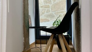 Un intérieur moderne avec un mur en pierre, une chaise noire aux pieds en bois et une vue par la fenêtre sur un bâtiment en pierre.
