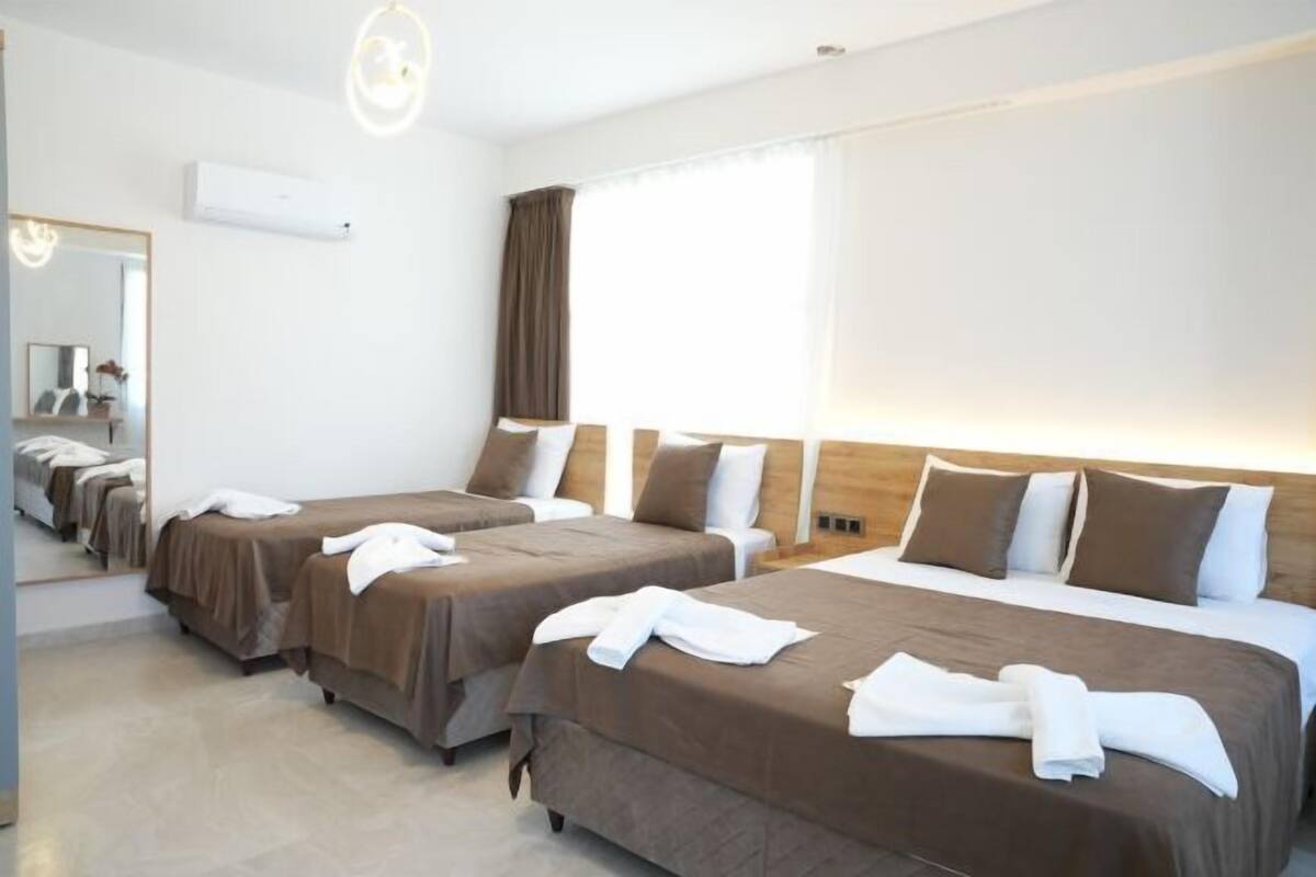 A hotel room with four single beds, a large window with curtains, a mirror, and a ceiling light fixture.
