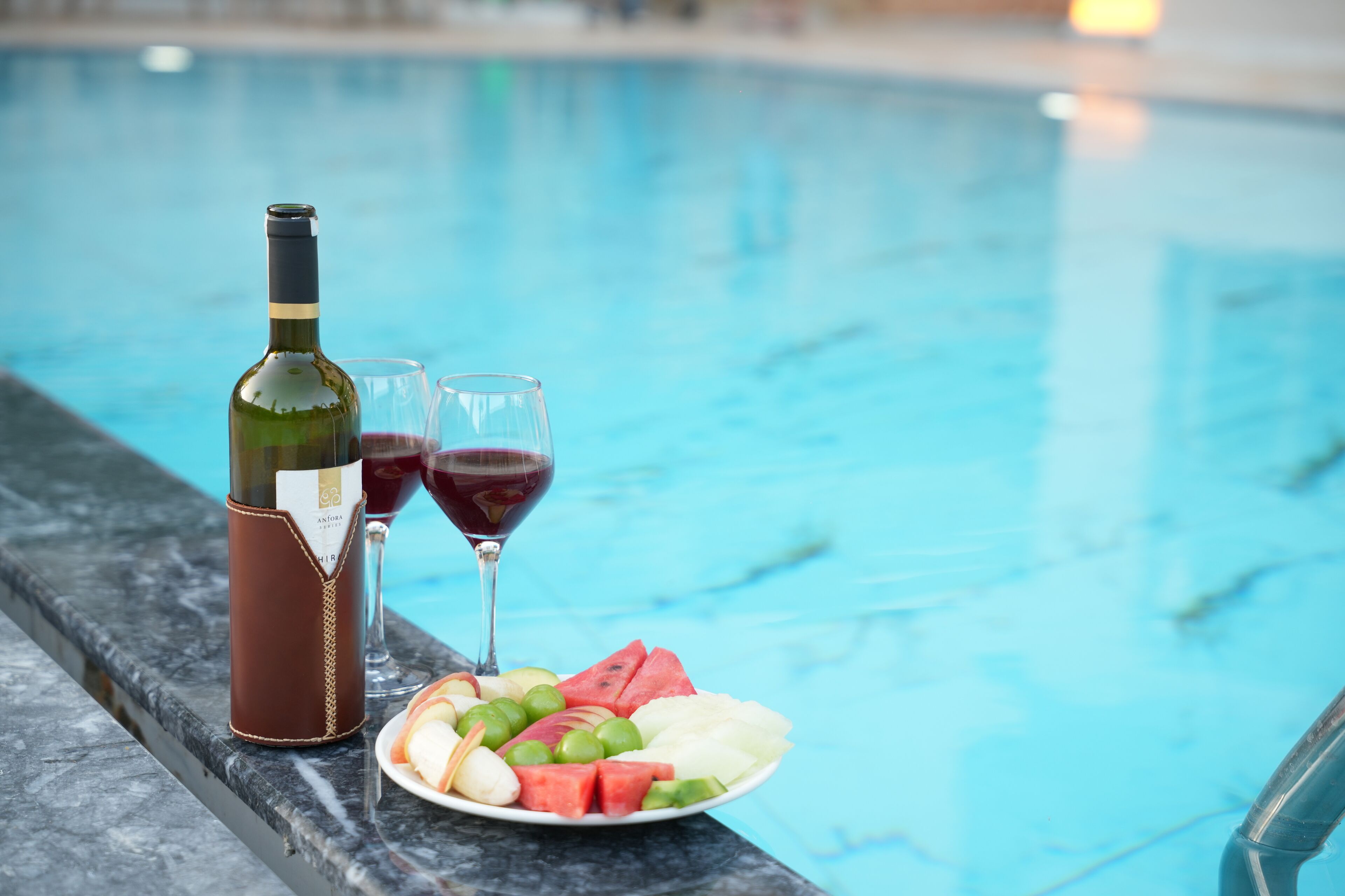 Une bouteille de vin, deux verres et une assiette de fruits près d’une piscine.