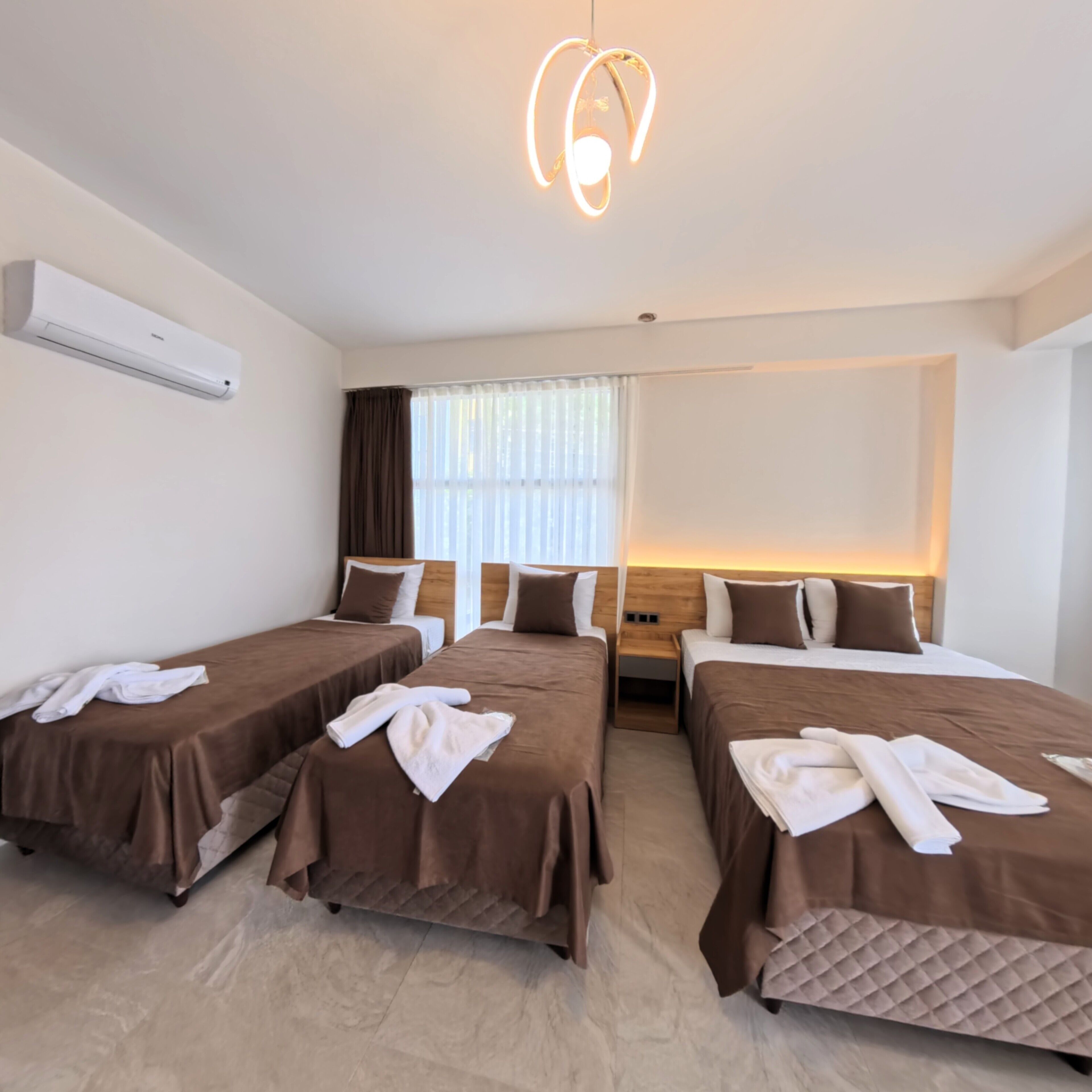 A hotel room with four single beds, each with a brown bedspread and white towels. There is a ceiling light fixture, a window with curtains, and a wall-mounted air conditioner.