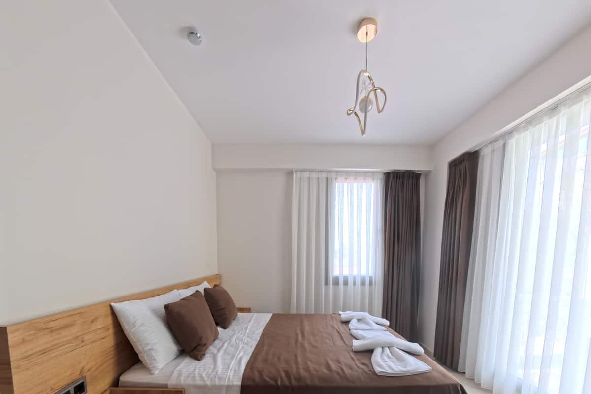 A modern hotel room with a large bed, a wooden bedside table, a built-in heater, and a view of the city through sheer curtains.