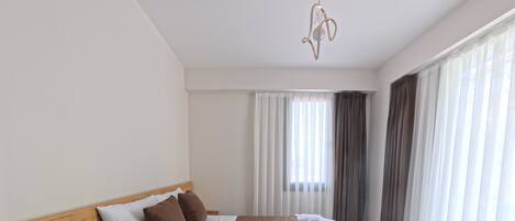Habitación de hotel moderna con una cama grande, una mesita de noche de madera, un calentador incorporado y vistas de la ciudad a través de cortinas traslúcidas.