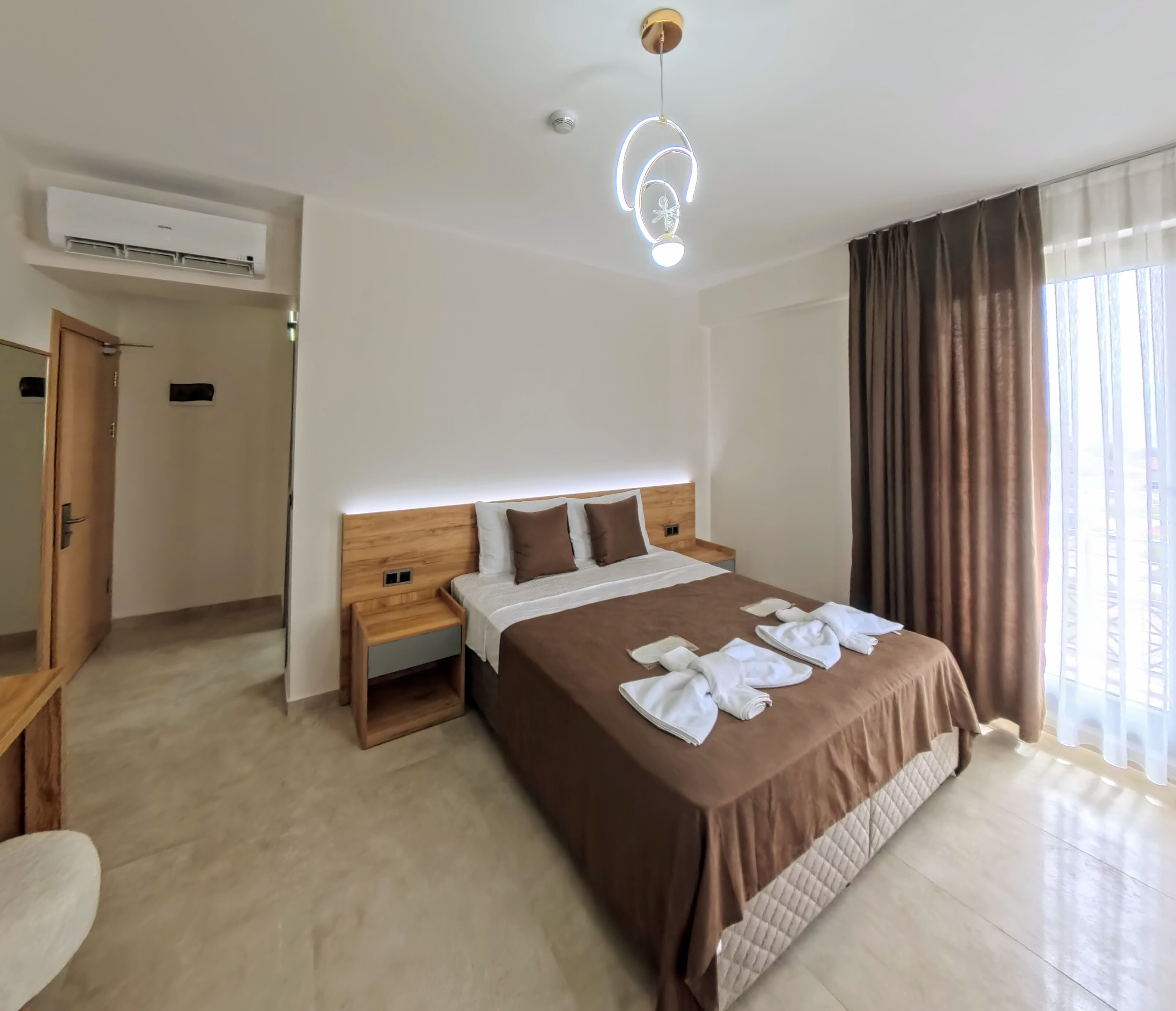 A hotel room with a large bed, a wooden headboard, a bedside table, an air conditioning unit, and a window with curtains.