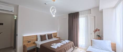 A hotel room with a large bed, a smaller bed, a desk, and a chair. There is a window with curtains and a plant on the desk.