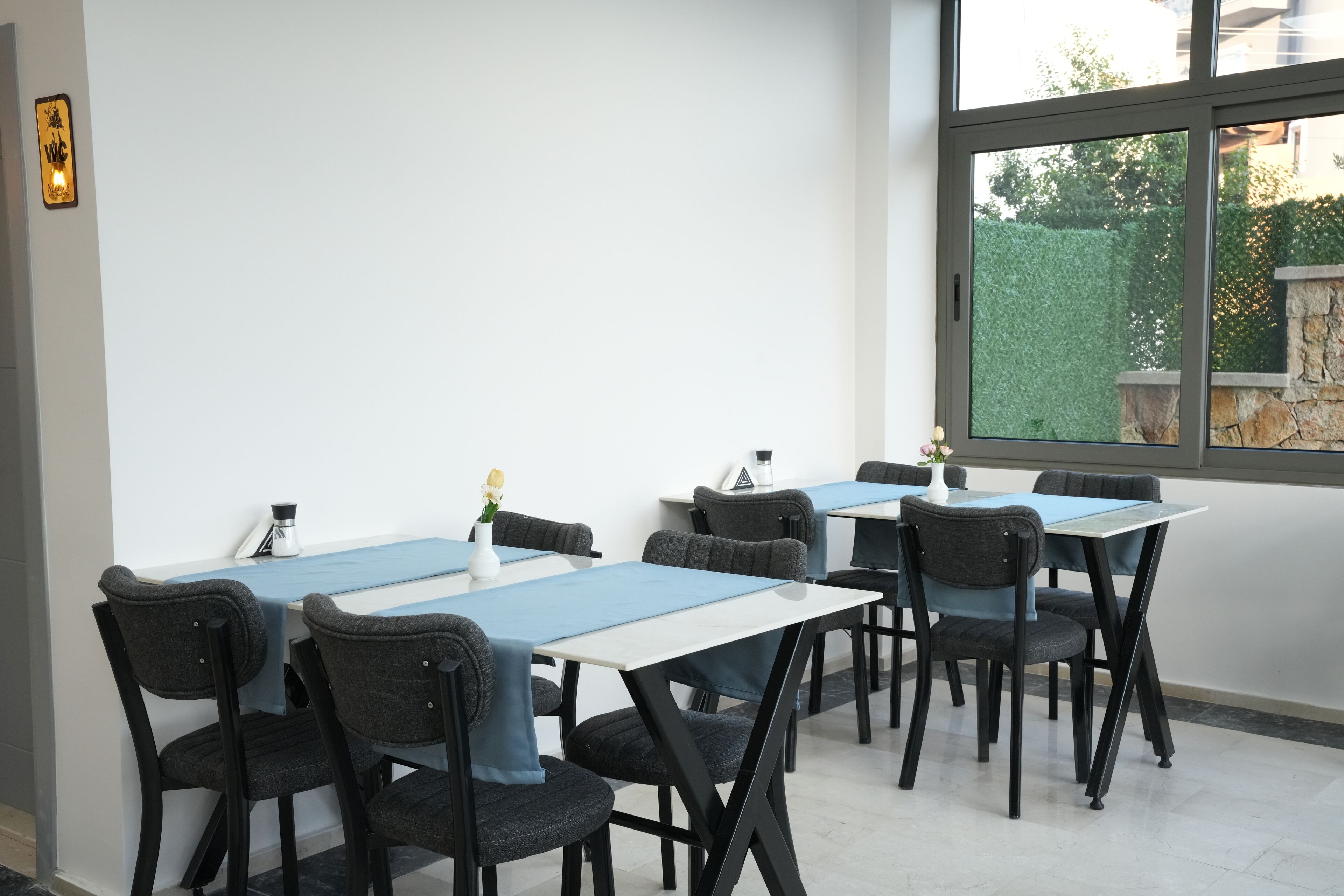 Un comedor con mesas y sillas, un ventanal con vistas a zonas verdes, y un aplique de pared.