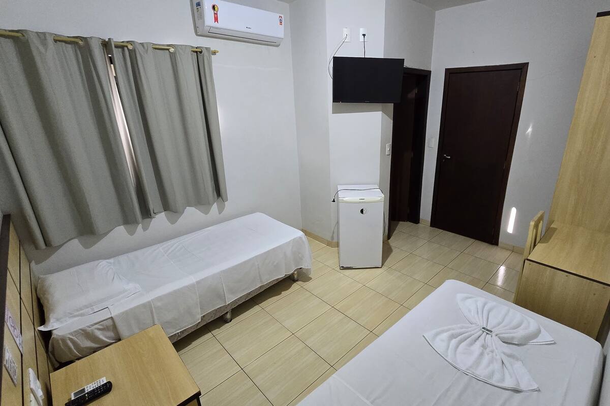 A small, clean room with a bed, a bedside table, a wardrobe, a small heater, and a TV mounted on the wall.