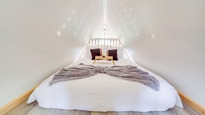 Ein Schlafzimmer mit schrägverdachter Decke, einem Bett mit weißer Bettdecke, zwei Kissen, einem Nachttisch aus Holz und einem kleinen Nachttisch mit Tablett.