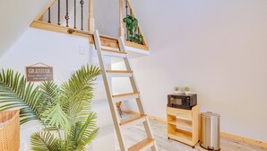 Eine Holzleiter führt zu einem erhöhten Bereich mit einem Pflanzenregal, einer Mikrowelle und einem Mülleimer in einem Zimmer mit Holzboden und weißer Wand.