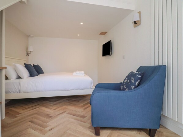 A bedroom with a bed, a blue armchair, a television, and a wooden herringbone floor. - Damson Tree Cottage (Bridport)