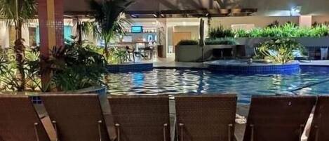 Un espace aménagé au bord de la piscine, avec des chaises longues et une piscine, le tout entouré de verdure et avec un bâtiment en arrière-plan.