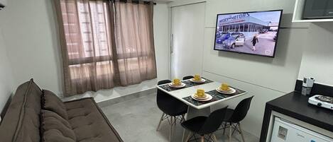 Un espace de vie compact comprenant un canapé, une table à manger avec des chaises, une petite kitchenette et un téléviseur fixé au mur.