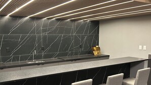 A modern bar area with a black marble backsplash, wooden bar counter, and three grey upholstered bar stools. - Melhores Flats - Seu refúgio (João Pessoa)