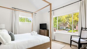 A modern hotel room with a four-poster bed, a flat-screen TV, a wooden nightstand, and a view of the outdoors. - Renovated! Coastal 3 Bedroom in the Heart of MTP (Mount Pleasant)