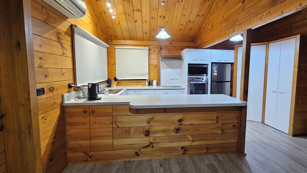 A wooden cabin interior with a kitchen featuring a central island, built-in cabinets, and a refrigerator. - Experience nature at it's finest! (Pukekohe)