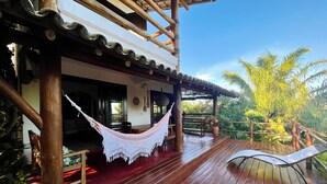 A wooden deck with a hammock and a lounge chair, surrounded by lush greenery and a clear blue sky. - Casa da Primavera (Porto Seguro)