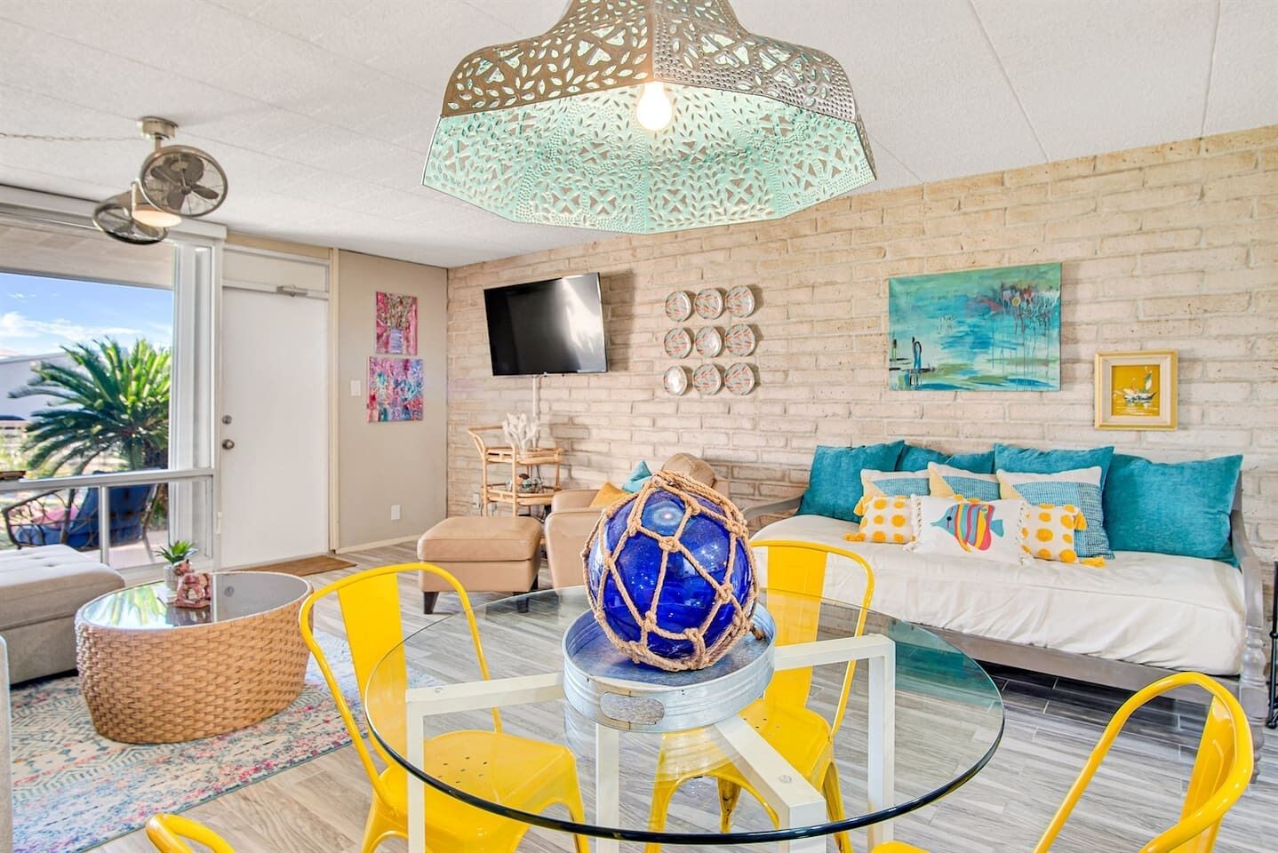 Un salon moderne avec une table basse en verre, des fauteuils jaunes, une boule décorative bleue, un téléviseur et un mur en briques.
