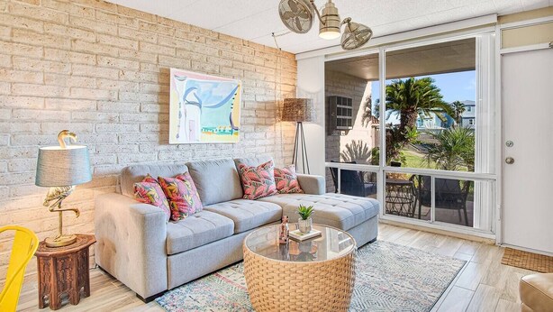 A living room with a grey sofa, colorful cushions, a wicker coffee table, a lamp, and a large window with a view of palm trees. - Condo in Beachfront Community! 4th nt FREE! (Port Aransas)