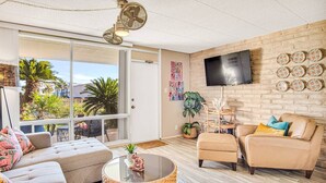 A modern living room with a sofa, armchair, and coffee table, a TV mounted on a brick wall, and a balcony with plants and a view. - Condo in Beachfront Community! 4th nt FREE! (Port Aransas)