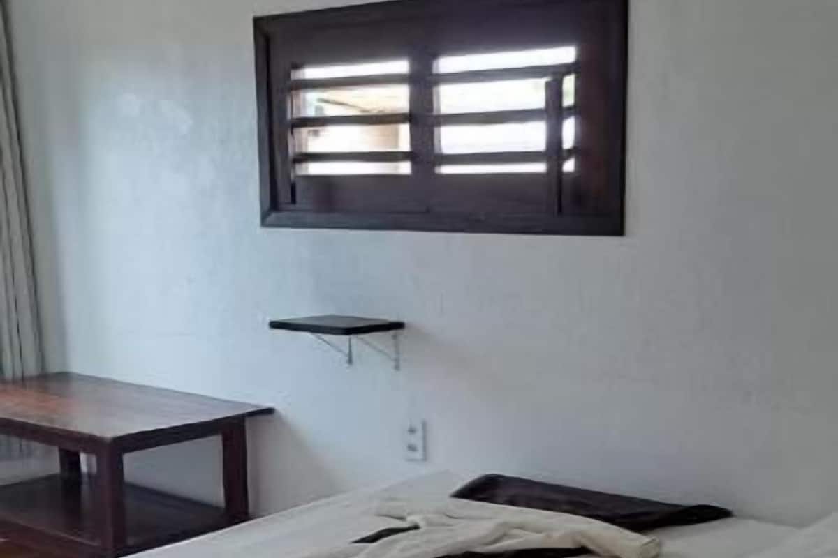 A single bed with a white mattress and dark bedding, a small table, a window with blinds, and a blue mat on the floor.