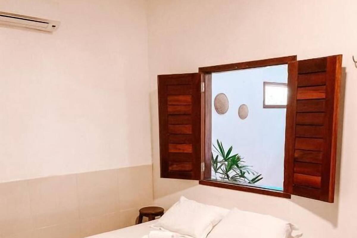 A neatly made bed with white linens and a red throw blanket, a small wooden nightstand, and a wall-mounted mirror with wooden shutters.