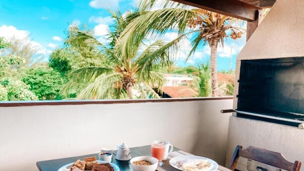 A balcony with a table set for breakfast, including plates of food, a crocheted item, and a view of palm trees and a house. - Vai Dá Certo Pousada  (Jijoca de Jericoacoara)