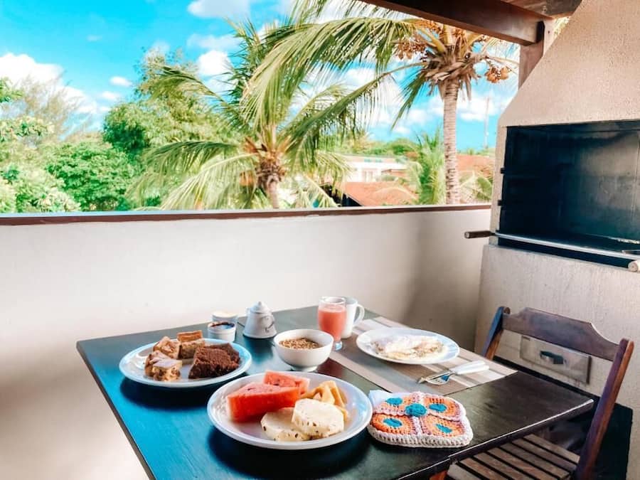 Un balcón con una mesa dispuesta para el desayuno, que incluye platos de comida, un adorno de crochet y vistas a palmeras y una casa.